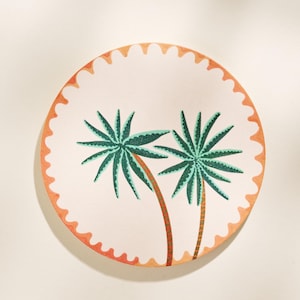 May include: Round decorative plate with a palm tree design. The plate features two green palm trees with brown trunks on a white background. The rim has an orange scalloped edge.