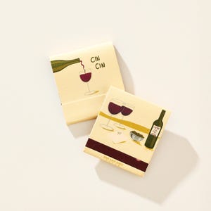 May include: Two square matchboxes with cream-colored covers. One features a green wine bottle pouring red wine into a glass with the text CIN CIN. The other shows two wine glasses, a bottle, a bowl of olives, and a napkin with the text ONE AND ONLY PAPER.