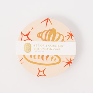 May include: Set of 4 coasters with a light pink background and a gold design of a croissant and a baguette. The text "SET OF 4 COASTERS good for hundreds of uses! made in the usa" is on a white label.