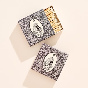 May include: Two square matchboxes with a dark gray background and white floral illustrations. Each box features a central oval containing a house drawing and the text Home Sweet Home. One box is open, revealing wooden matches with white tips.