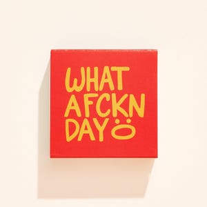 May include: A square red box featuring the yellow text WHAT AFCKN DAY with a smiley face symbol at the end. The lettering is in a casual, hand-drawn style, centered on the front face of the box, which has a flat, matte finish and sharp edges.