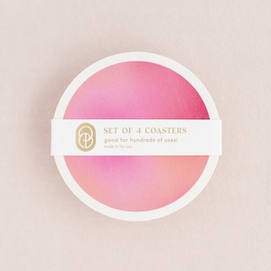 May include: A set of four pink and white round coasters with a gradient design. The coasters have a white border and a gold logo with the text "SET OF 4 COASTERS good for hundreds of uses! made in the usa".