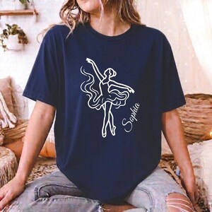 Op de afbeelding: Een marineblauw T-shirt met korte mouwen en een witte lijntekening van een ballerina in een zwierige jurk. De naam Sophia is in een cursief lettertype aan de rechterkant van de danseres gedrukt. Het T-shirt heeft een ronde hals en een losse pasvorm.