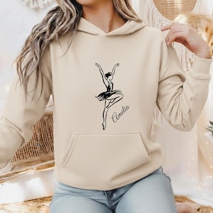 Op de afbeelding: Een beige hoodie met een zwarte grafische print van een ballerina in een danshouding. Het ontwerp bevat de naam Amelia in een cursief lettertype onder de danseres. Het kledingstuk heeft lange mouwen en een kangoeroezak aan de voorzijde.