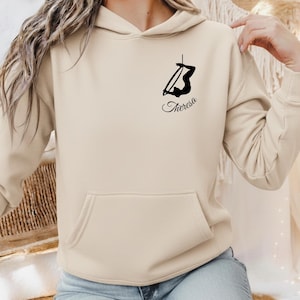 May include: A beige hoodie with a front pocket and a drawstring hood. The hoodie features a black graphic of a person on a hoop, with the name "Theresa" in cursive below the graphic. The hoodie is made of a soft material.