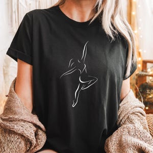 Puede incluir: Una camiseta negra de manga corta con un gráfico de líneas blancas que representa a una persona bailando en una pose dinámica. El diseño consiste en líneas blancas simples y continuas que perfilan la silueta de una persona en pleno movimiento.
