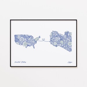 May include: A framed print featuring blue patterned maps of the United States and Libya, connected by a heart-shaped line. The words "United States" and "Libya" are written below the maps in a dark blue font. The print is set against a white background.