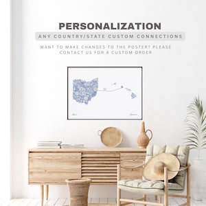 May include: A framed art print featuring a blue and white floral design of the Ohio and Hawaii state outlines connected by a line with a heart. The print is displayed on a white wall above a wooden cabinet.