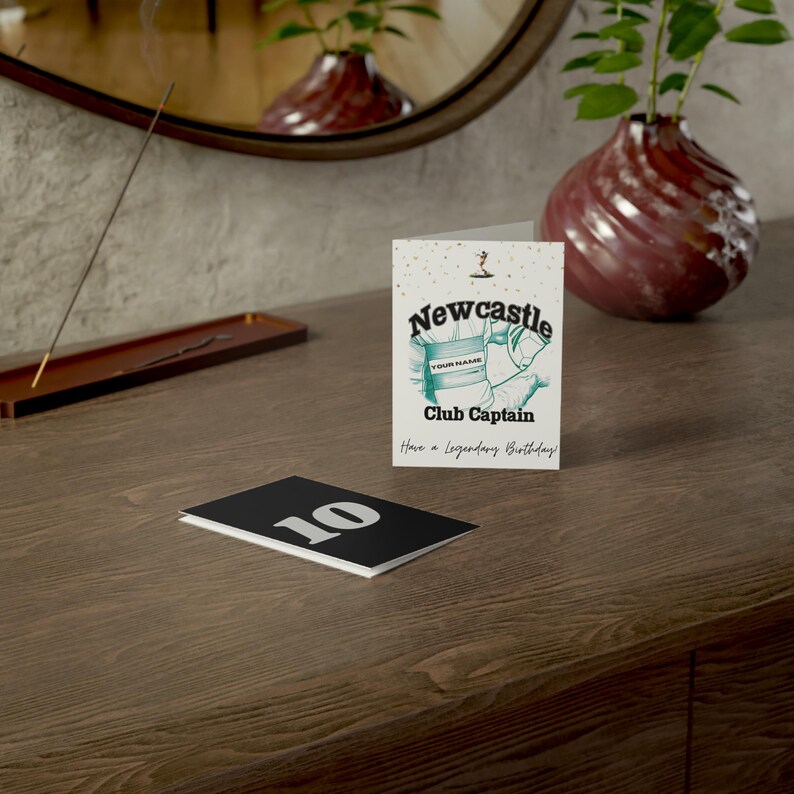 May include: A greeting card with the word "Newcastle" in a stylized font, with a teal and white illustration. The card reads "Club Captain" and "Have a Legendary Birthday!". A black card with the number "10" is also visible.