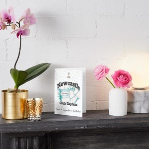 May include: A still life arrangement featuring a greeting card with the text "Newcastle Club Captain," a potted orchid, a decorative candle holder, a small vase with pink roses, and a modern lamp with a marble base.