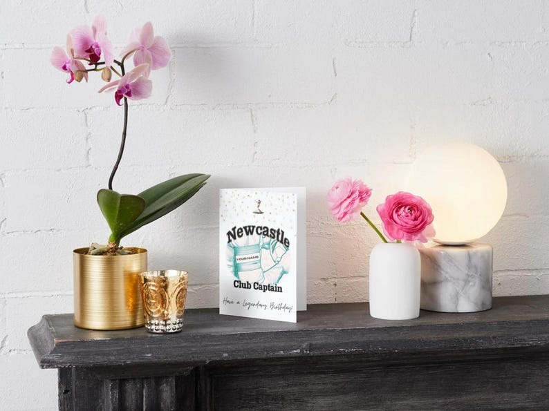 May include: A still life arrangement featuring a greeting card with the text "Newcastle Club Captain," a potted orchid, a decorative candle holder, a small vase with pink roses, and a modern lamp with a marble base.