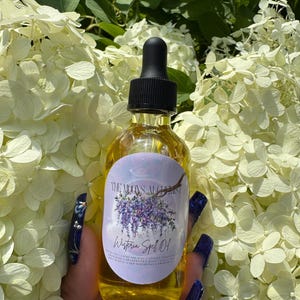 Könnte beinhalten: Eine klare Glasflasche mit schwarzem Tropfverschluss, gefüllt mit einer goldenen Flüssigkeit. Die Flasche hat ein Etikett mit dem Text "The Moon's Alchemist" und "Wisteria Spell 01" mit einem lila Blumendesign. Die Flasche wird vor weißen Blumen gehalten.