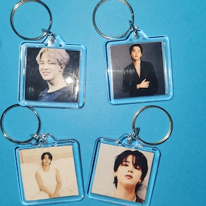 May include: Four clear acrylic keychains with silver key rings. Each keychain has a square photo of a person. The keychains have a light blue border and are arranged on a light blue background.
