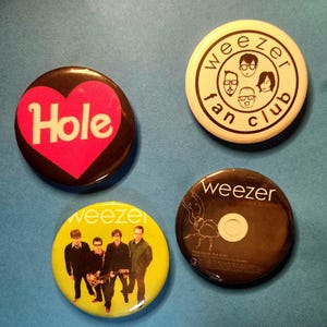 Può includere: Quattro bottoni rotondi con vari disegni. Un bottone ha un cuore rosa con la scritta "Hole" in bianco. Un altro è bianco con "Weezer fan club" e facce. Il terzo è giallo con una foto della band. L'ultimo è nero con uno scarabeo e "Weezer".