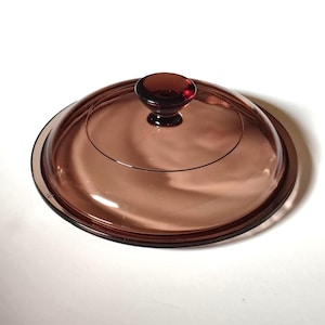 May include: A clear, round, amber-colored glass lid with a matching knob. The lid has a dark brown rim and a circular design etched into the glass. The knob is a darker shade of amber.