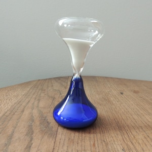 May include: An hourglass with a clear glass top and a deep blue base. White sand is flowing from the top chamber to the bottom. The hourglass is sitting on a wooden surface.