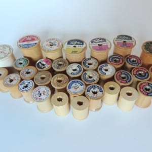 May include: A collection of vintage wooden spools of thread, featuring various labels with text and colorful designs. The spools are arranged on a white surface, showcasing their aged appearance and diverse branding.