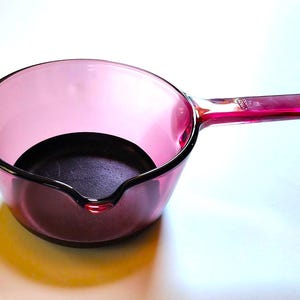 May include: A vintage pink glass saucepan with a handle. The pan has a rounded shape with a pouring spout and a dark interior. The handle is a solid piece of glass, and the pan is set against a white background.