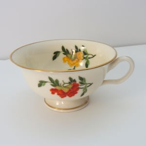 May include: Cream-colored teacup with a gold rim and base, featuring floral designs in red and yellow with green leaves. The cup has a curved handle and is set against a white background.