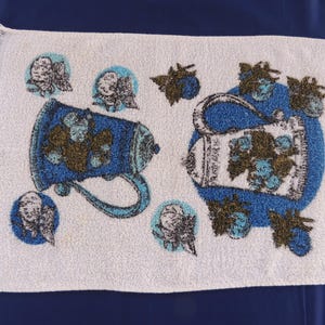 May include: White kitchen towel with a blue and green design of two pitchers with flowers and leaves. The towel has a fringed edge.