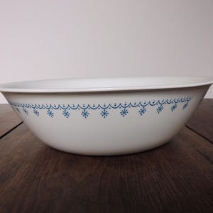 May include: White ceramic bowl with a blue snowflake pattern around the rim. The bowl has a wide, shallow shape and sits on a dark wooden surface. A decorative kitchenware item.