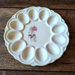 May include: A round, off-white ceramic deviled egg platter featuring a scalloped edge and twelve oval-shaped indentations. The center of the plate is decorated with a small, detailed illustration of a single pink rose with green leaves.