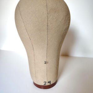 May include: A beige canvas-covered head form for millinery or wig display. The mannequin head features vertical stitched seams and the number 24 printed in black ink on the front and near the base. It sits on a circular brown wooden base.