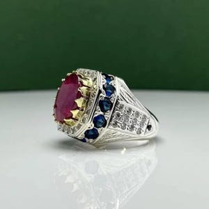 Puede incluir: Un anillo de plata con una gran piedra roja de corte ovalado engastada en un bisel dorado. La banda está decorada con una hilera de zafiros azules redondos y piedras claras más pequeñas, con detalles metálicos grabados en los laterales.