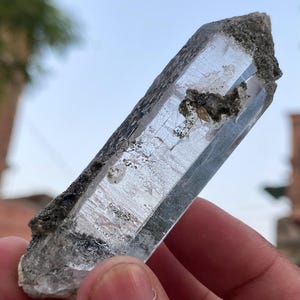 May include: A clear quartz crystal with a rough, dark gray base. The crystal is elongated and has visible internal structures. The light reflects off the facets, creating a shimmering effect. The crystal is held in a hand.