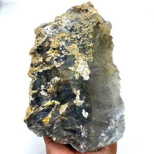 May include: A large, raw crystal formation with a mix of colors. The stone features areas of dark gray, black, and yellow, contrasted by lighter, translucent sections. The surface has a rough, textured appearance, with small crystalline formations.