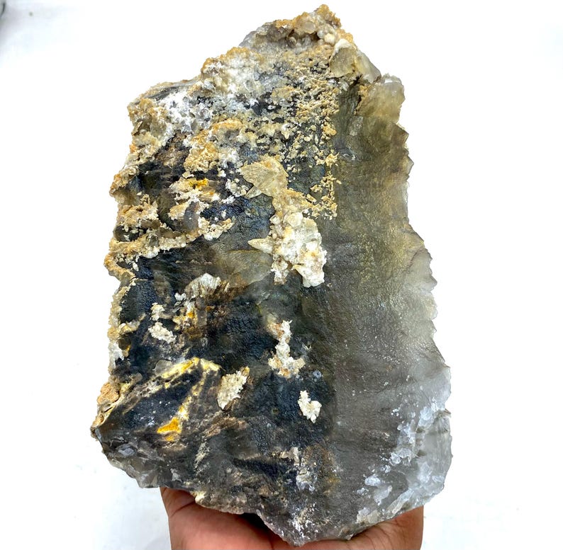 May include: A large, raw crystal formation with a mix of colors. The stone features areas of dark gray, black, and yellow, contrasted by lighter, translucent sections. The surface has a rough, textured appearance, with small crystalline formations.