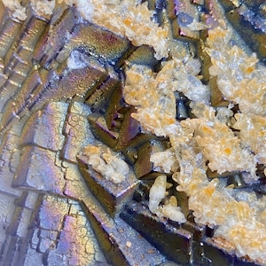 May include: Close-up of a mineral formation with a metallic, iridescent sheen. The surface displays a geometric pattern of layered, angular structures in shades of silver, purple, and gold. Clusters of crystalline formations are scattered across the surface.