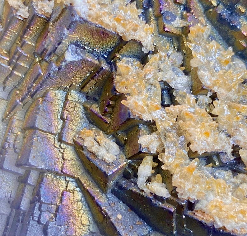 May include: Close-up of a mineral formation with a metallic, iridescent sheen. The surface displays a geometric pattern of layered, angular structures in shades of silver, purple, and gold. Clusters of crystalline formations are scattered across the surface.