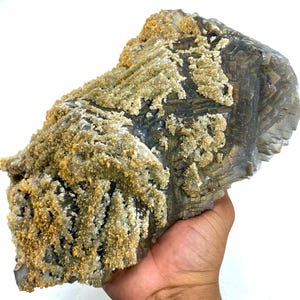 May include: A large, rough-hewn mineral specimen with a layered, geometric structure. The stone has a dark, metallic gray base color, with areas of iridescent sheen. The surface is partially covered with a light tan, crystalline coating, creating a textured appearance.