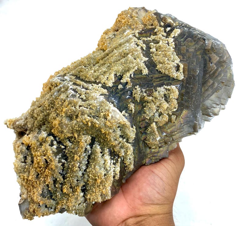 May include: A large, rough-hewn mineral specimen with a layered, geometric structure. The stone has a dark, metallic gray base color, with areas of iridescent sheen. The surface is partially covered with a light tan, crystalline coating, creating a textured appearance.