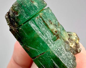 Puede incluir: Un cristal de esmeralda en bruto y alargado que presenta un tono verde intenso con estrías verticales visibles, texturas superficiales naturales e inclusiones minerales oscuras. La piedra preciosa muestra una calidad translúcida.