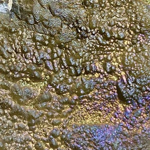 May include: Close-up of a textured surface with a metallic sheen. The surface has a bumpy, irregular texture with a mix of colors, including gold, green, purple, and blue, creating an iridescent effect. The overall appearance is organic and abstract.