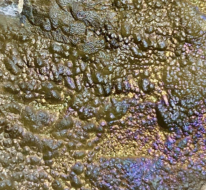 May include: Close-up of a textured surface with a metallic sheen. The surface has a bumpy, irregular texture with a mix of colors, including gold, green, purple, and blue, creating an iridescent effect. The overall appearance is organic and abstract.