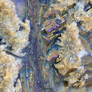 May include: Close-up of a mineral specimen with iridescent, metallic surfaces in shades of blue, purple, and gold. The crystalline structure is visible, with clusters of small, beige crystals. The texture is rough and detailed.