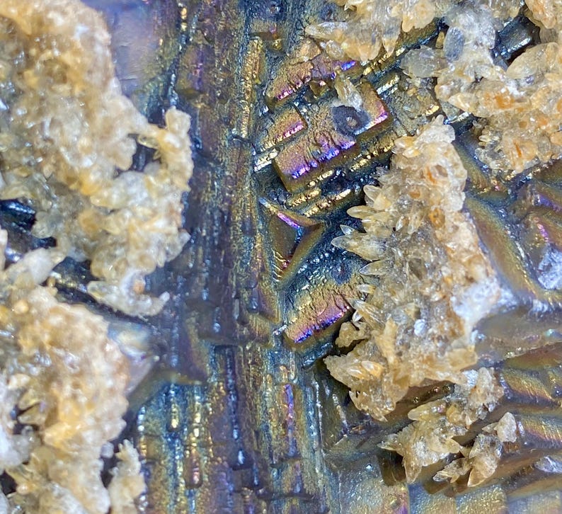 May include: Close-up of a mineral specimen with iridescent, metallic surfaces in shades of blue, purple, and gold. The crystalline structure is visible, with clusters of small, beige crystals. The texture is rough and detailed.