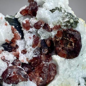 Könnte beinhalten: Ein Mineralexemplar mit tiefroten Granatkristallen, die in eine Matrix aus weißem Quarz und durchscheinenden kristallinen Strukturen eingebettet sind. Die Gruppe zeigt dunkle, kantige Granatformationen neben metallisch grünen Einschlüssen.