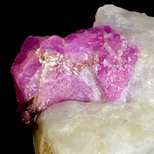 May include: A raw mineral specimen featuring a vibrant, translucent magenta crystal formation embedded within a larger, opaque white quartz matrix. The magenta section displays a jagged, crystalline texture with reflective facets and irregular edges.