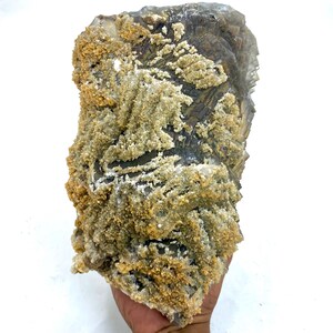 May include: A large, rough-hewn mineral specimen with a layered, crystalline structure. The rock features a mix of gray, tan, and white colors, with a textured, granular surface. The mineral is held in a hand against a white background.