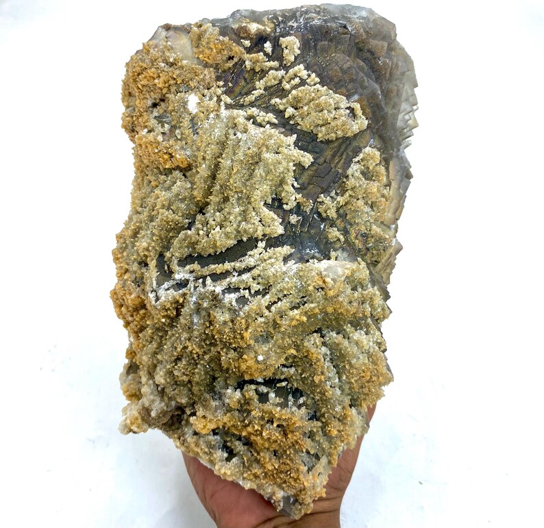 May include: A large, rough-hewn mineral specimen with a layered, crystalline structure. The rock features a mix of gray, tan, and white colors, with a textured, granular surface. The mineral is held in a hand against a white background.