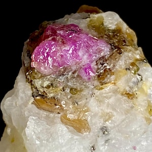 May include: A raw mineral specimen featuring a vibrant, translucent magenta-pink crystal cluster embedded within a textured, off-white quartz matrix. The central pink formation displays a crystalline structure with reflective facets and inclusions.