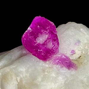 May include: A vibrant, deep magenta-colored crystal formation embedded within a textured, translucent white mineral matrix. The primary crystal features sharp, irregular facets and a glossy surface, contrasting against the opaque, rough white base.