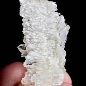 Puede incluir: Un grupo de formaciones minerales cristalinas claras. Los cristales están apilados en una estructura en capas, con una apariencia ligeramente translúcida. El mineral se sostiene sobre un fondo negro, resaltando su forma natural.