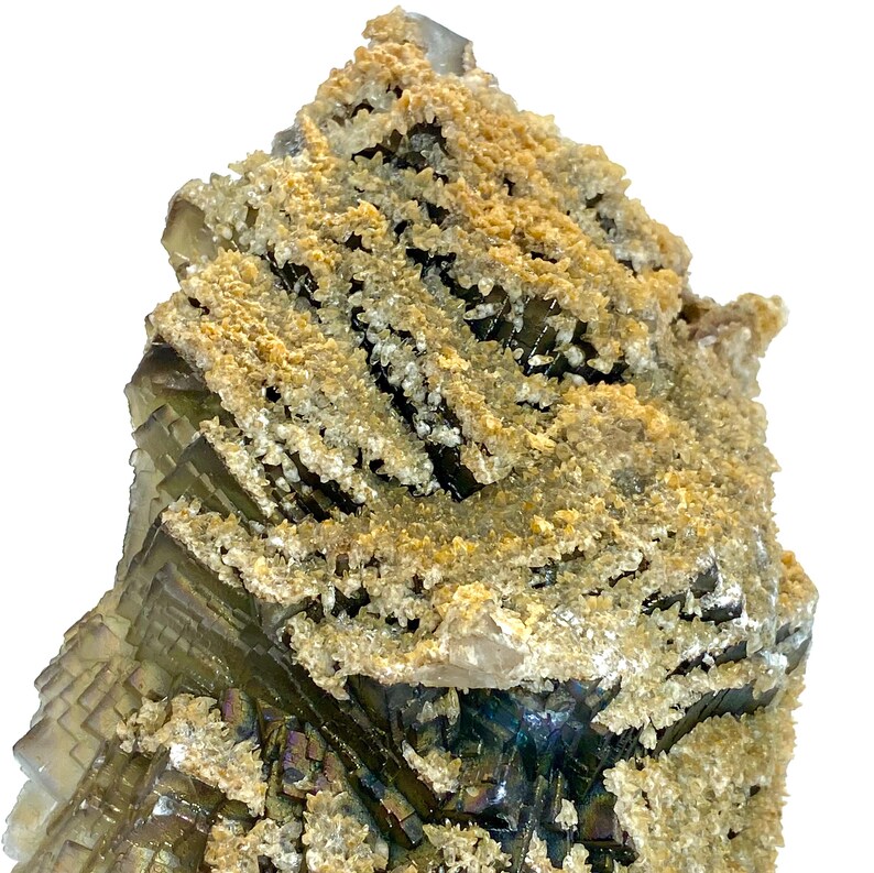 May include: A close-up of a natural mineral specimen. The mineral features layered, translucent green and purple sections with a crystalline structure. The surface is coated with a layer of tan and white granular material, creating a textured appearance.