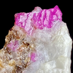 May include: A raw mineral specimen featuring a prominent, vibrant magenta-pink crystal formation at the top. The base consists of translucent white crystalline structures interspersed with patches of textured, earthy brown mineral deposits.