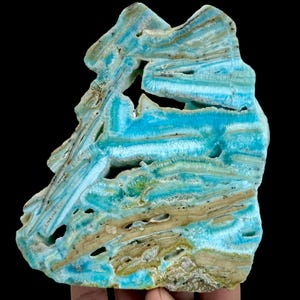 May include: A striking, irregularly shaped mineral specimen with a vibrant turquoise hue. The stone features layered bands of color, including shades of blue, tan, and hints of green. The surface has natural holes and a rough texture, showcasing its unique geological formation.